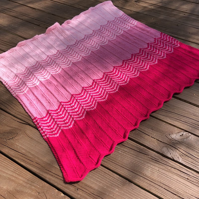 A chevron knit blanket in shades of pink (hot pink, medium pink, pale pink) on a wooden deck