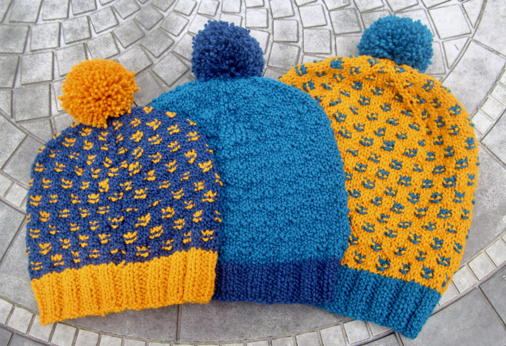 Three handknit hats sizes small to large, in shades of navy, turquoise and bright yellow, lay on a ceramic table.
