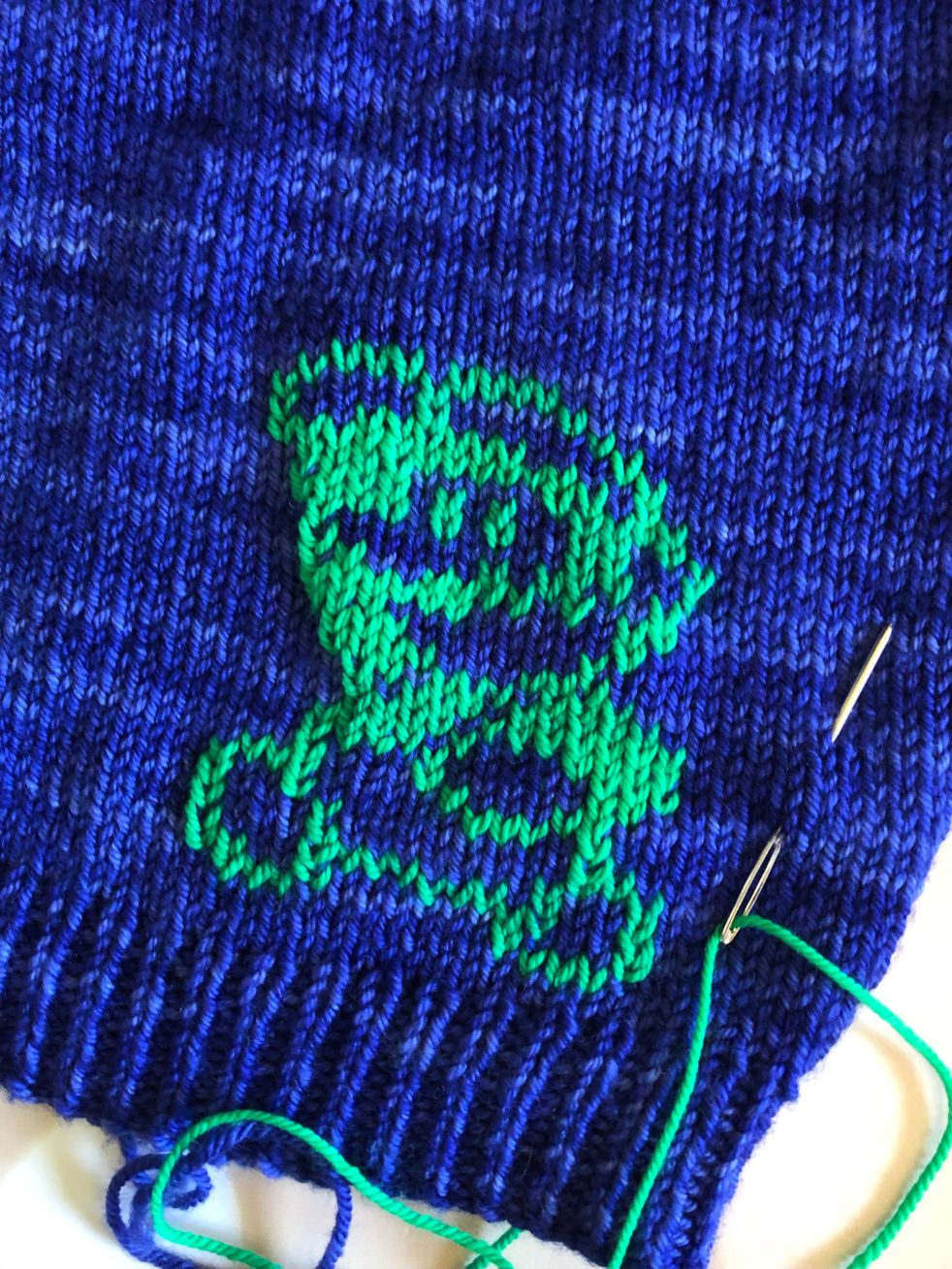 A photo of the hem of a royal blue sweater. Luigi of Super Mario Brothers has been duplicate stitched onto the sweater in bright green yarn. A needle threaded with green yarn is tucked into the stitches for more embroidery to come.