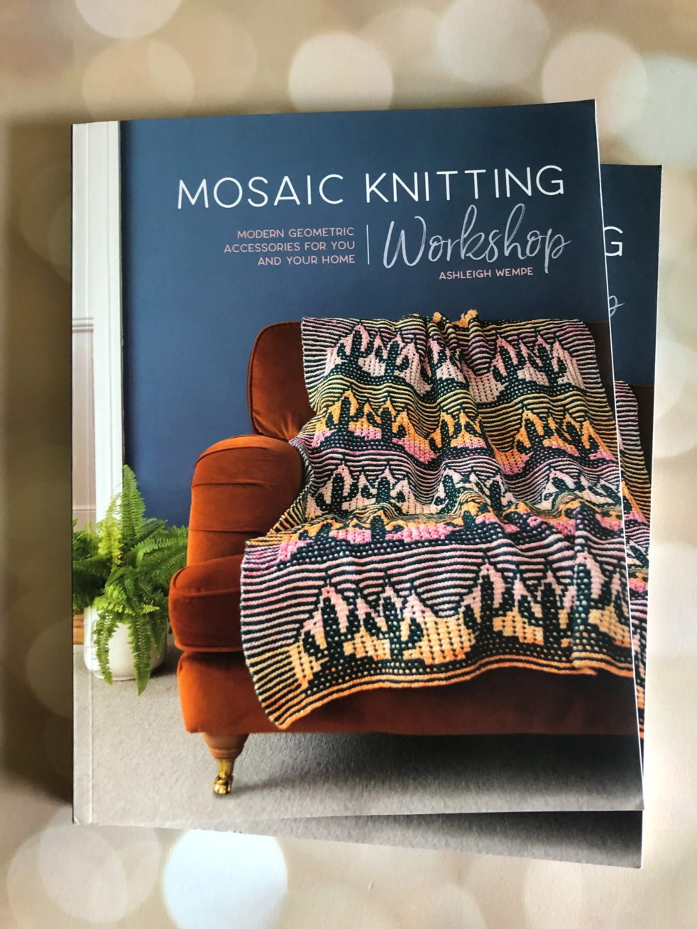 Two copies of Mosaic Knitting Workshop lay on a pink bokeh background. The cover features a mosaic knit blanket featuring cacti against a gradient background of pinks, oranges and yellows.