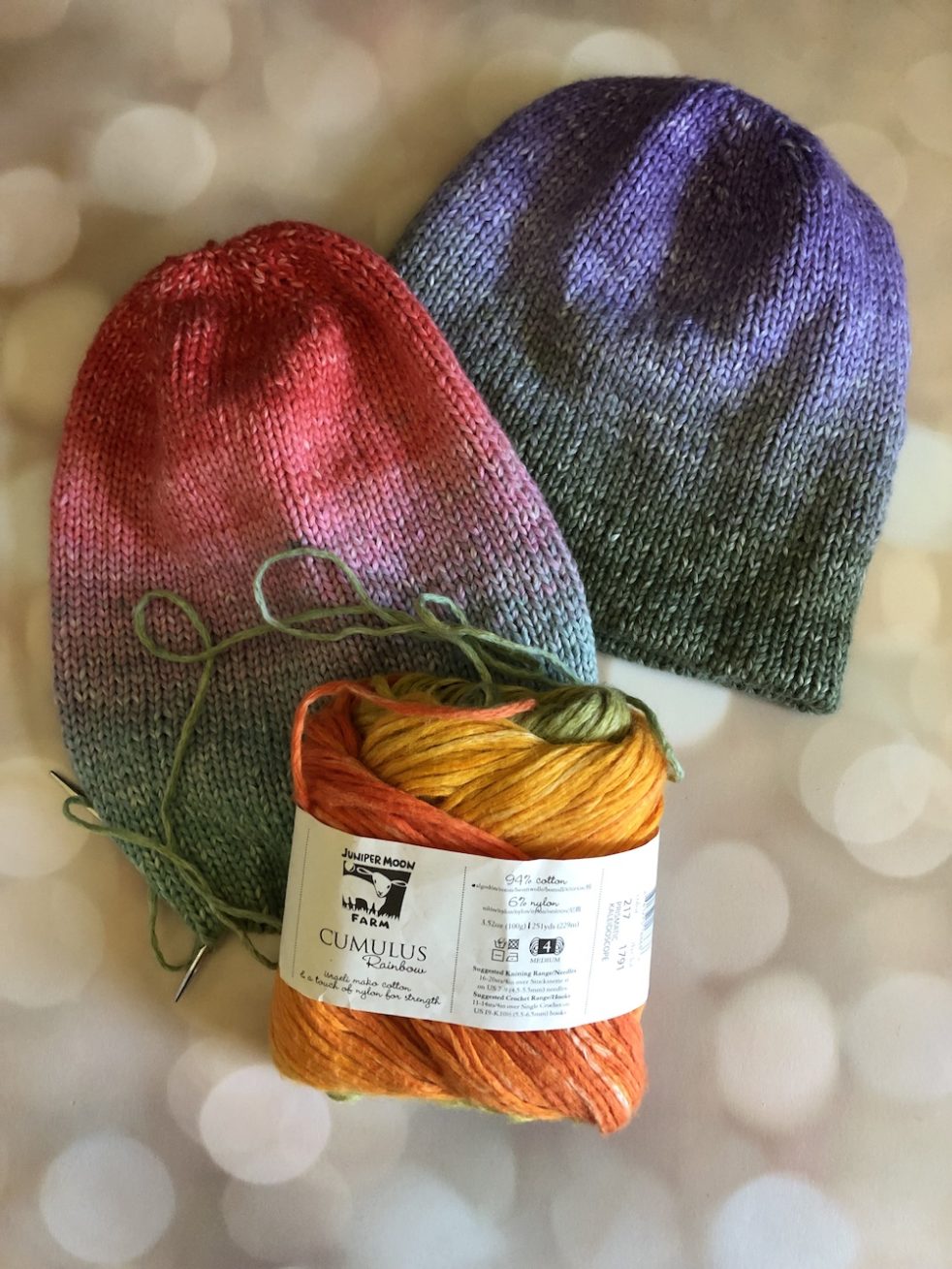 Two hats knit in gradient yarns. One in purple to green is already knit. One in red to green to orange is in progress on the needles.