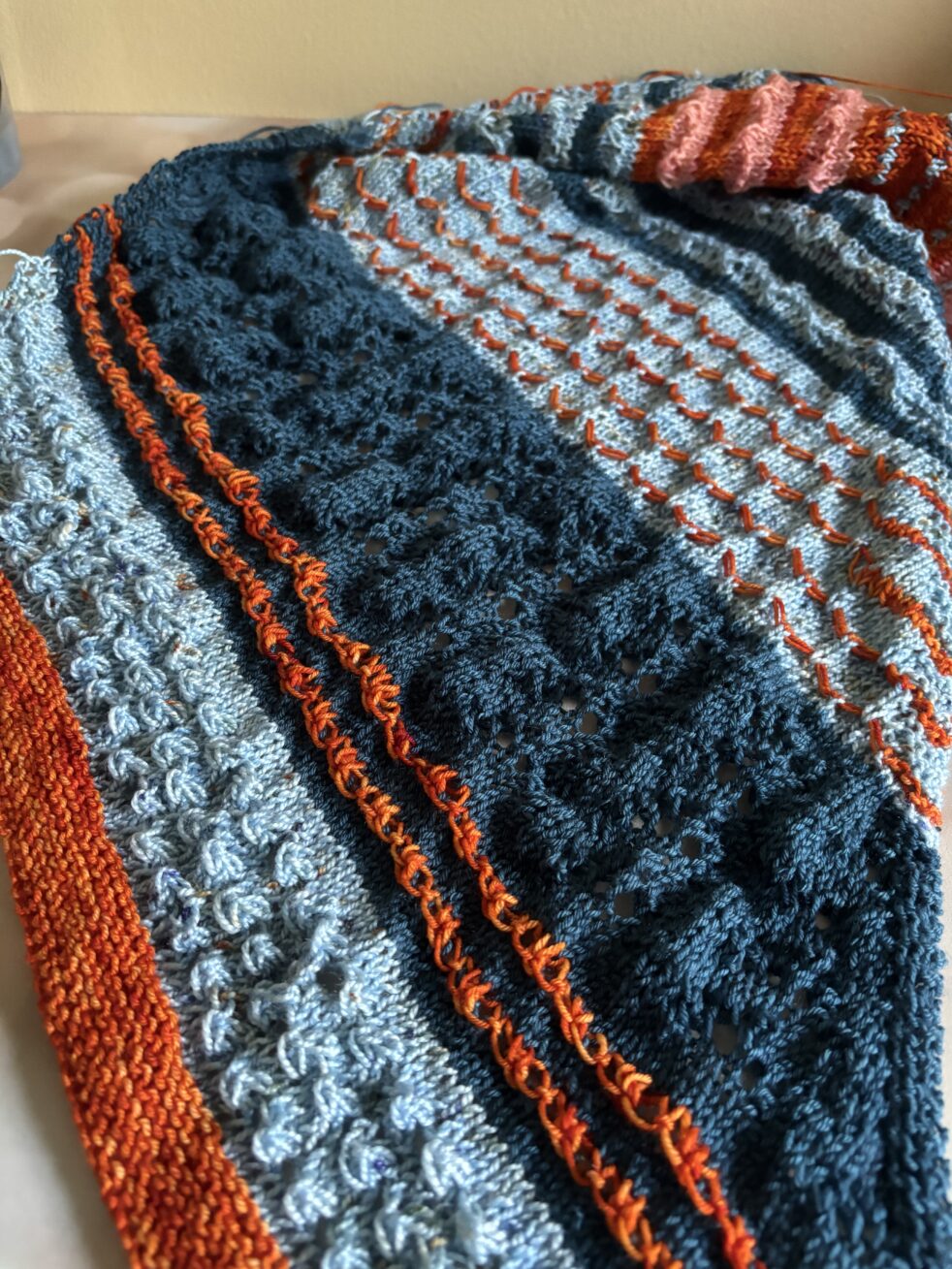 A sampler shawl knit in various patterns in two shades of orange and two shades of blue.